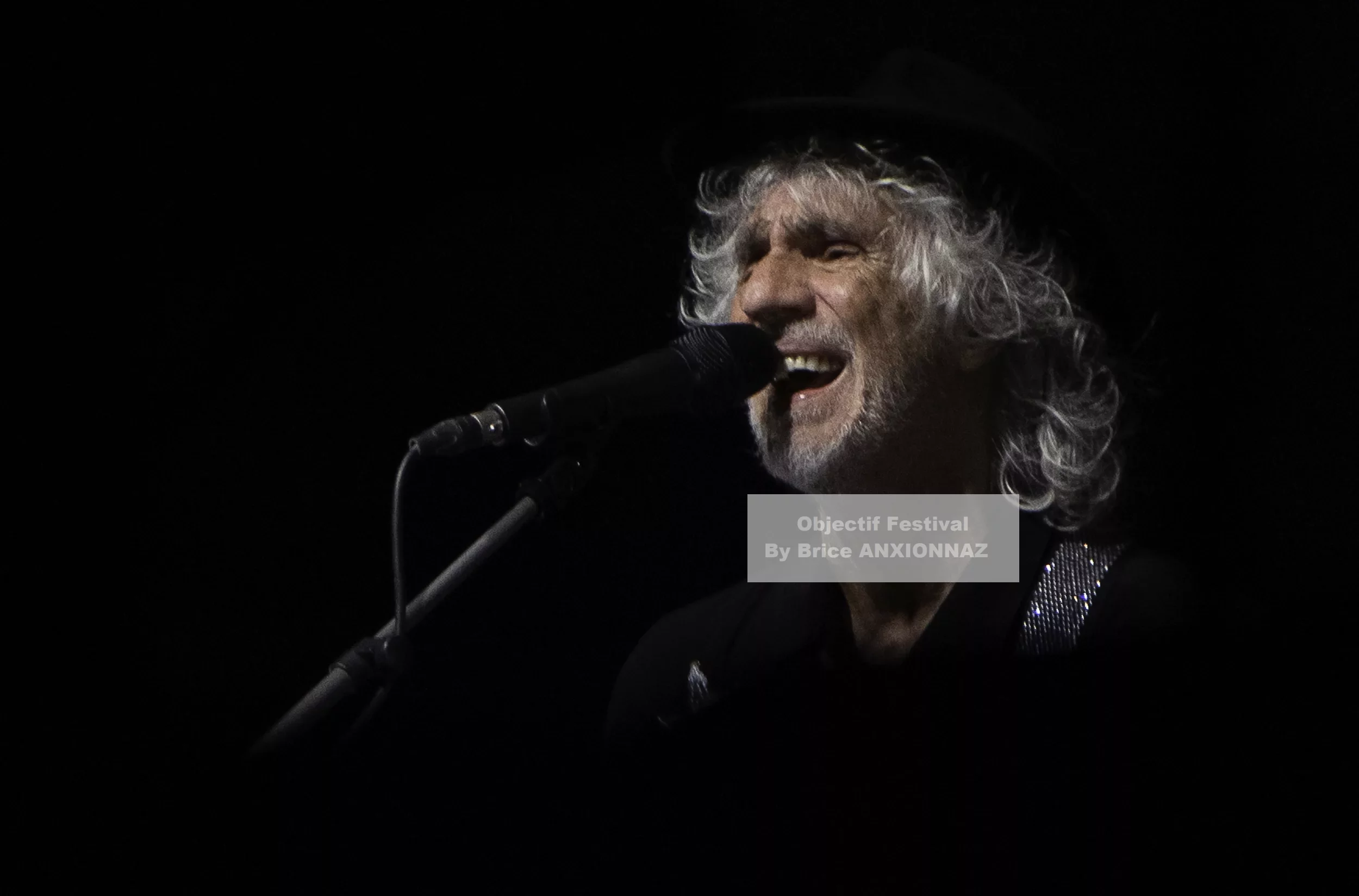 Louis Bertignac — Louis Bertignac — Un homme chante au microphone, éclairé par une lumière douce, avec des cheveux argentés et un chapeau noir, créant une ambiance intimiste sur scène.