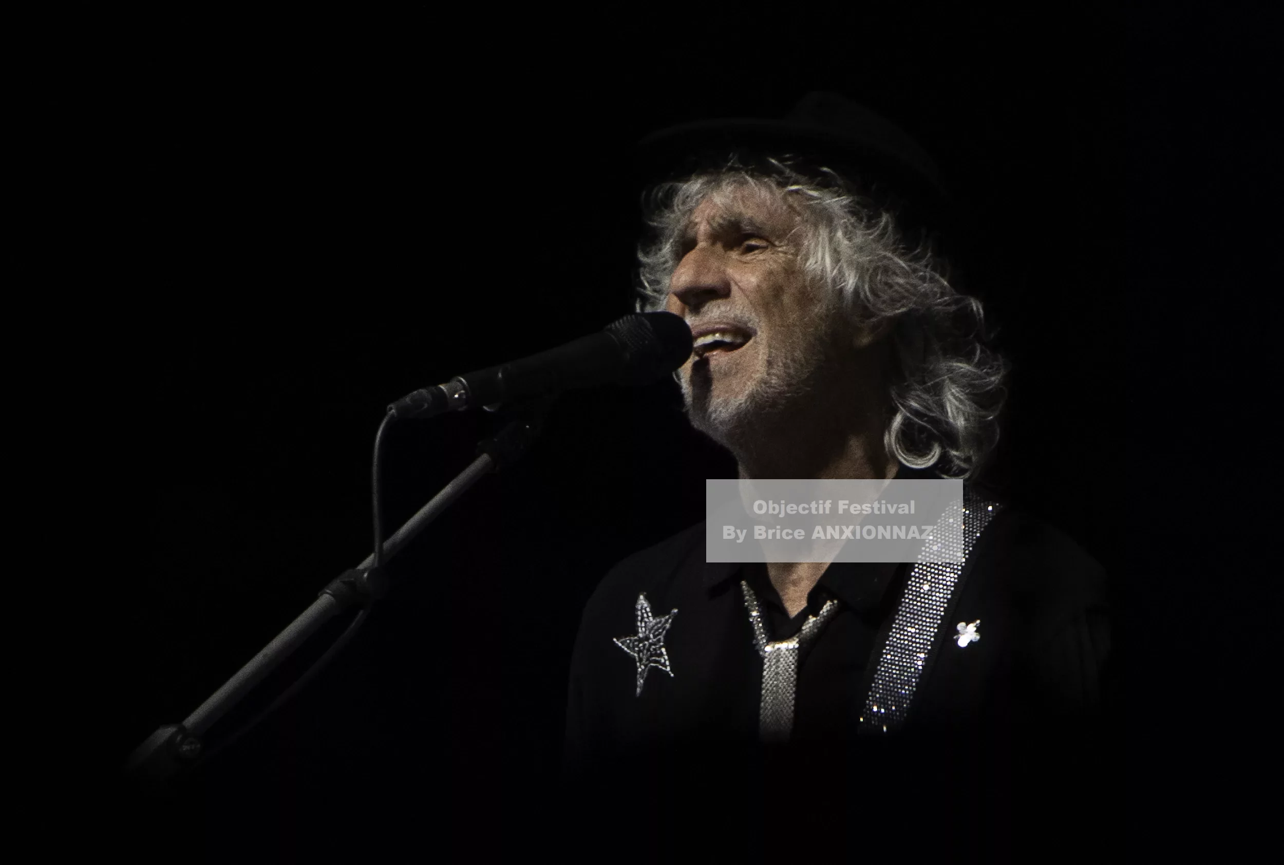 Louis Bertignac — Louis Bertignac — Un homme chante au microphone, vêtu d'une chemise noire ornée d'une étoile, avec des cheveux bouclés et argentés, dans une ambiance sombre et éclairée par un éclairage doux.