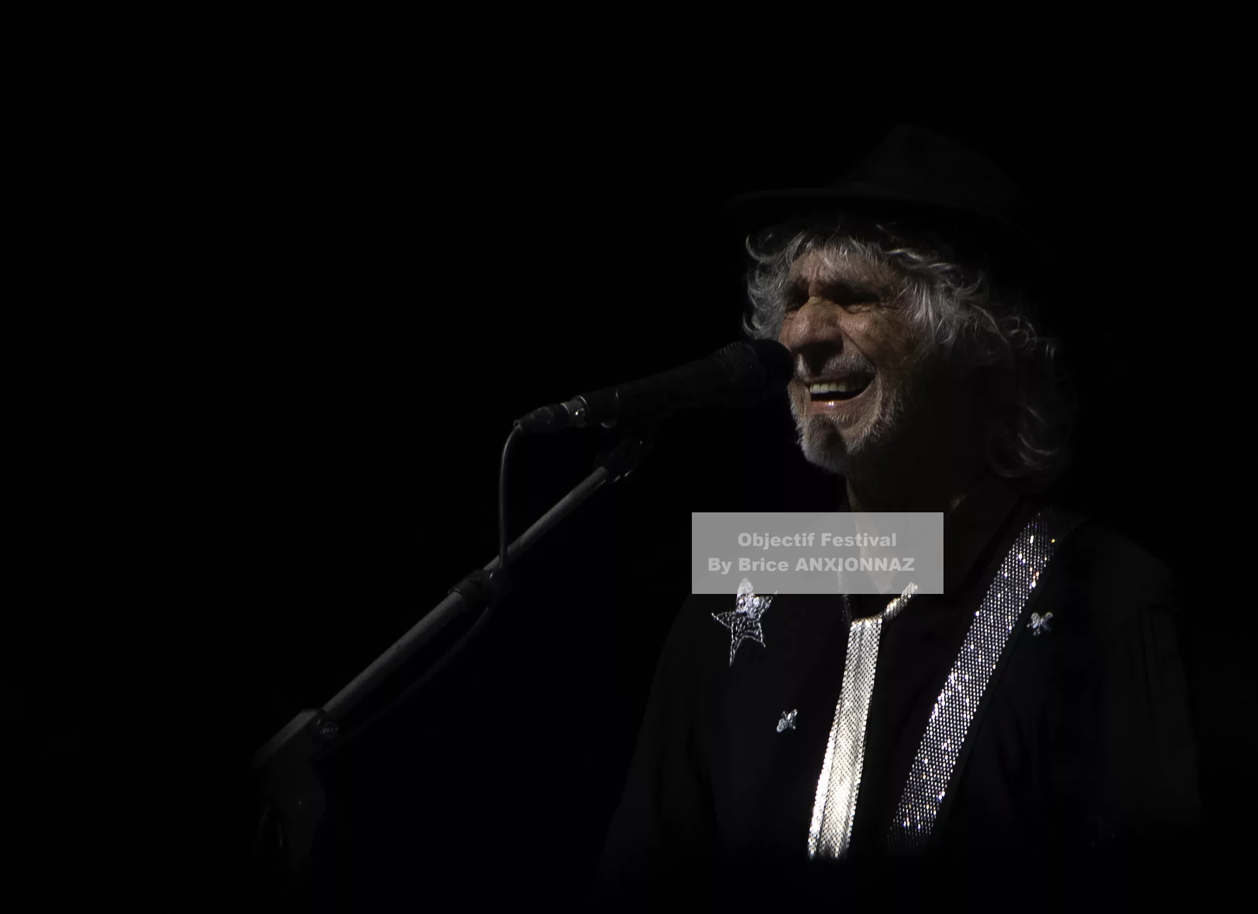Louis Bertignac — Un homme souriant chante au microphone, vêtu d'une chemise noire ornée de paillettes, sur fond sombre, créant une ambiance intimiste et captivante. 21 07 2026, Hyeres (21/07/2026)