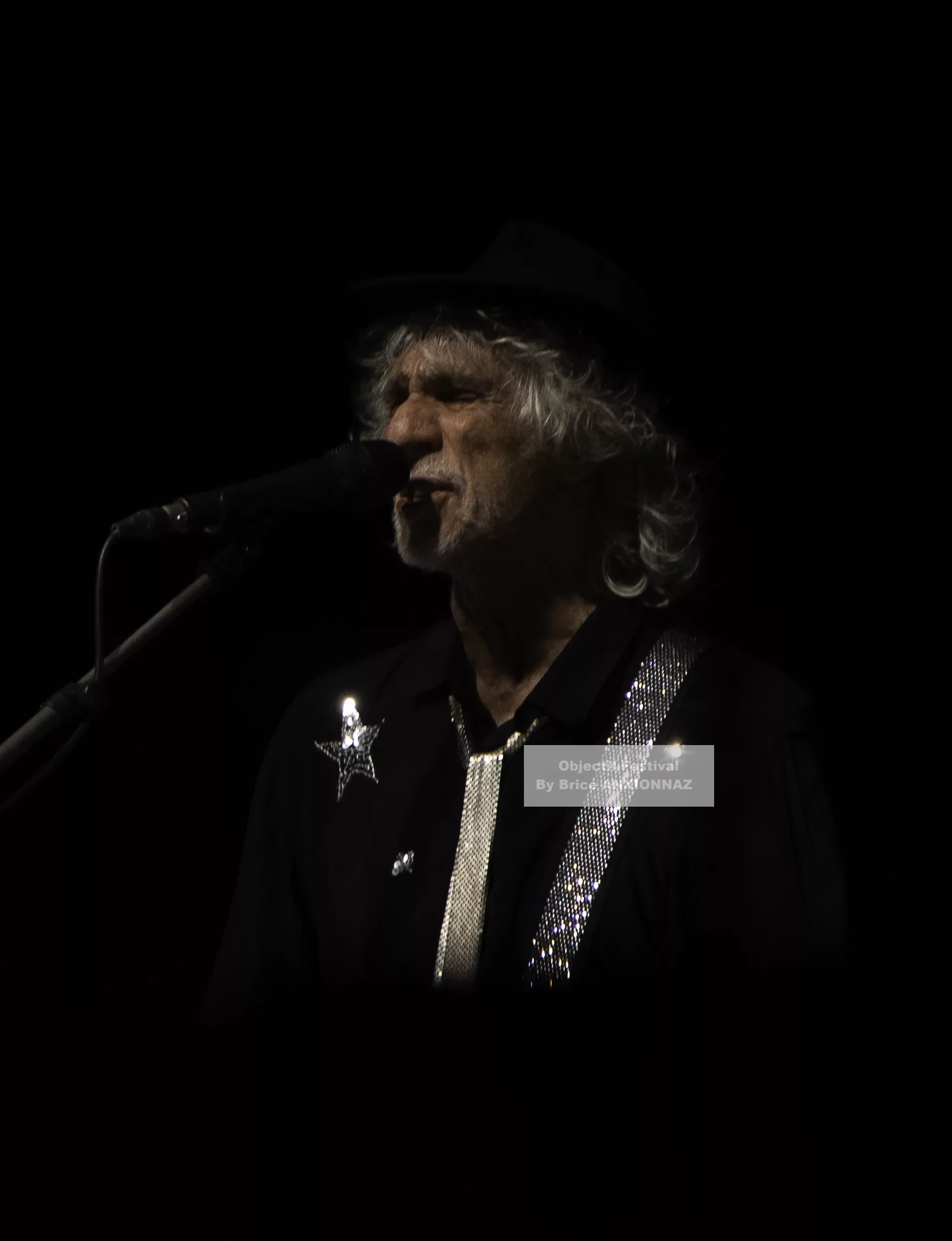 Louis Bertignac — Louis Bertignac — Un artiste chante au microphone, vêtu d'une chemise noire ornée de paillettes et d'un chapeau, dans une ambiance sombre avec un éclairage focalisé sur lui.