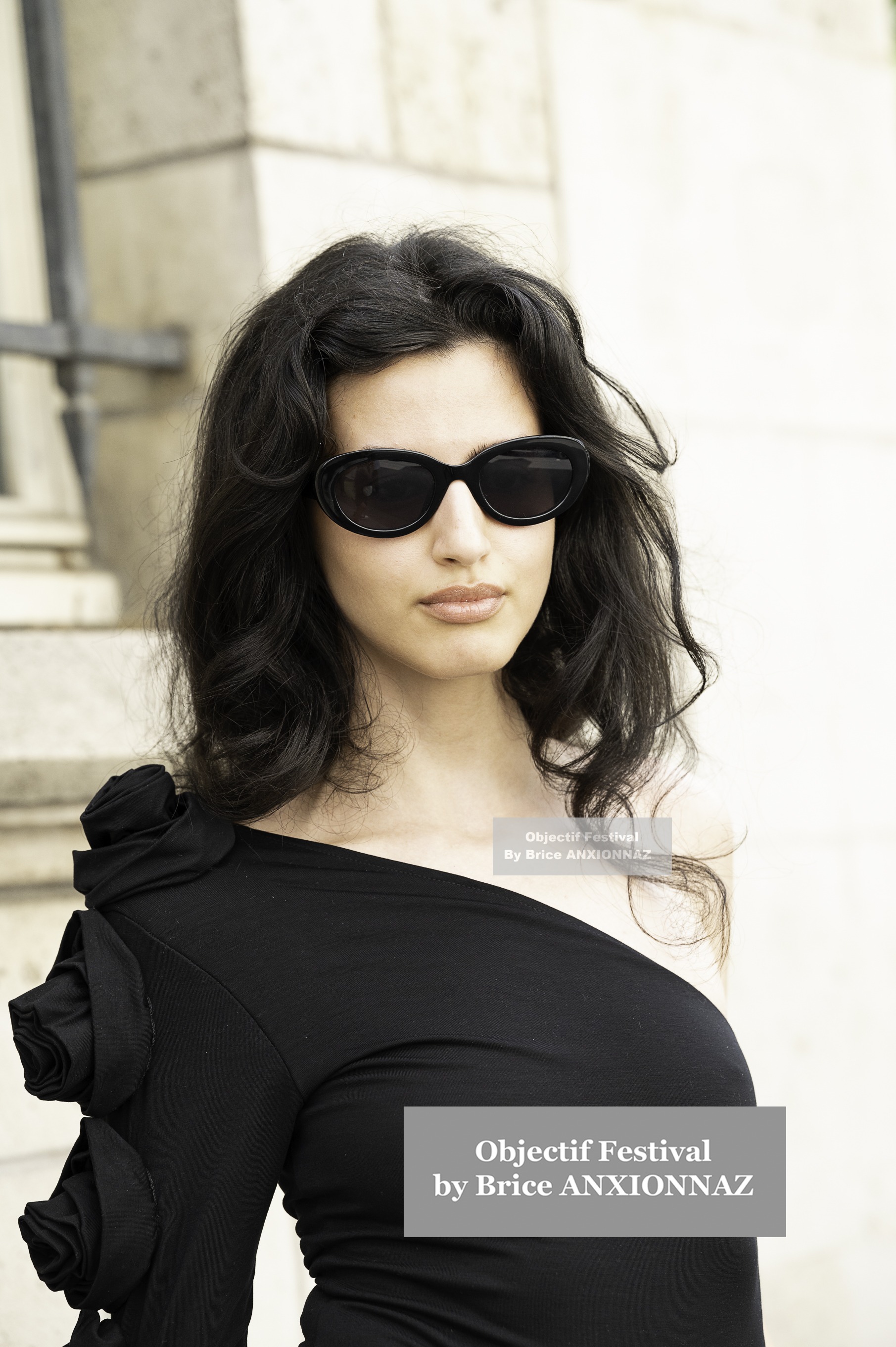 Christian Dior — Une personne porte un haut noir à une épaule avec des détails floraux, des lunettes de soleil, et se tient devant un mur en pierre, dans une lumière douce et naturelle. Pfw Haute Couture Fw25, Paris (24/06/2024)