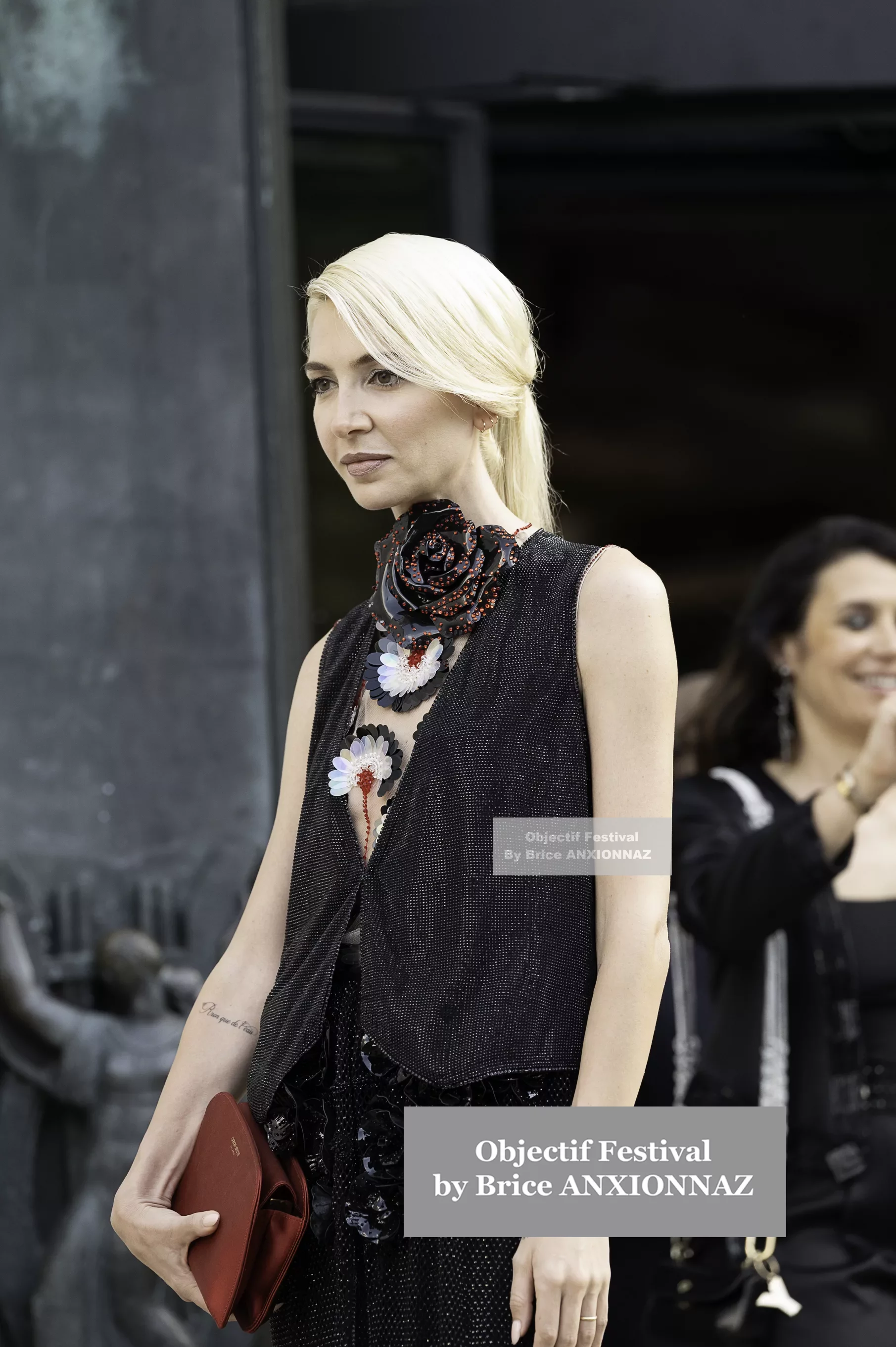 Giorgio Armani Prive — Une personne se tient debout, vêtue d'une tenue noire ornée de fleurs, avec un sac à main rouge, tandis qu'une autre personne souriante passe en arrière-plan. Pfw Haute Couture Fw25, Paris (27/06/2024)
