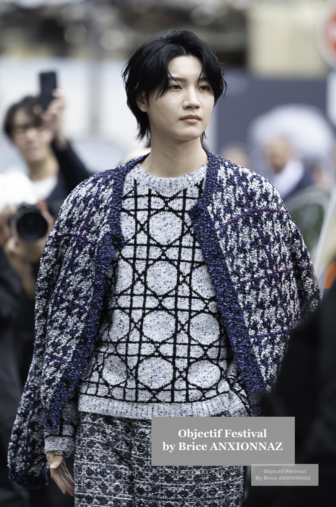 Dori Sakurada — Un individu marche avec assurance, vêtu d'un pull à motifs géométriques et d'une veste texturée, tandis que des photographes capturent le moment en arrière-plan. Pfw Menswear Ss25, Paris (21/06/2024)