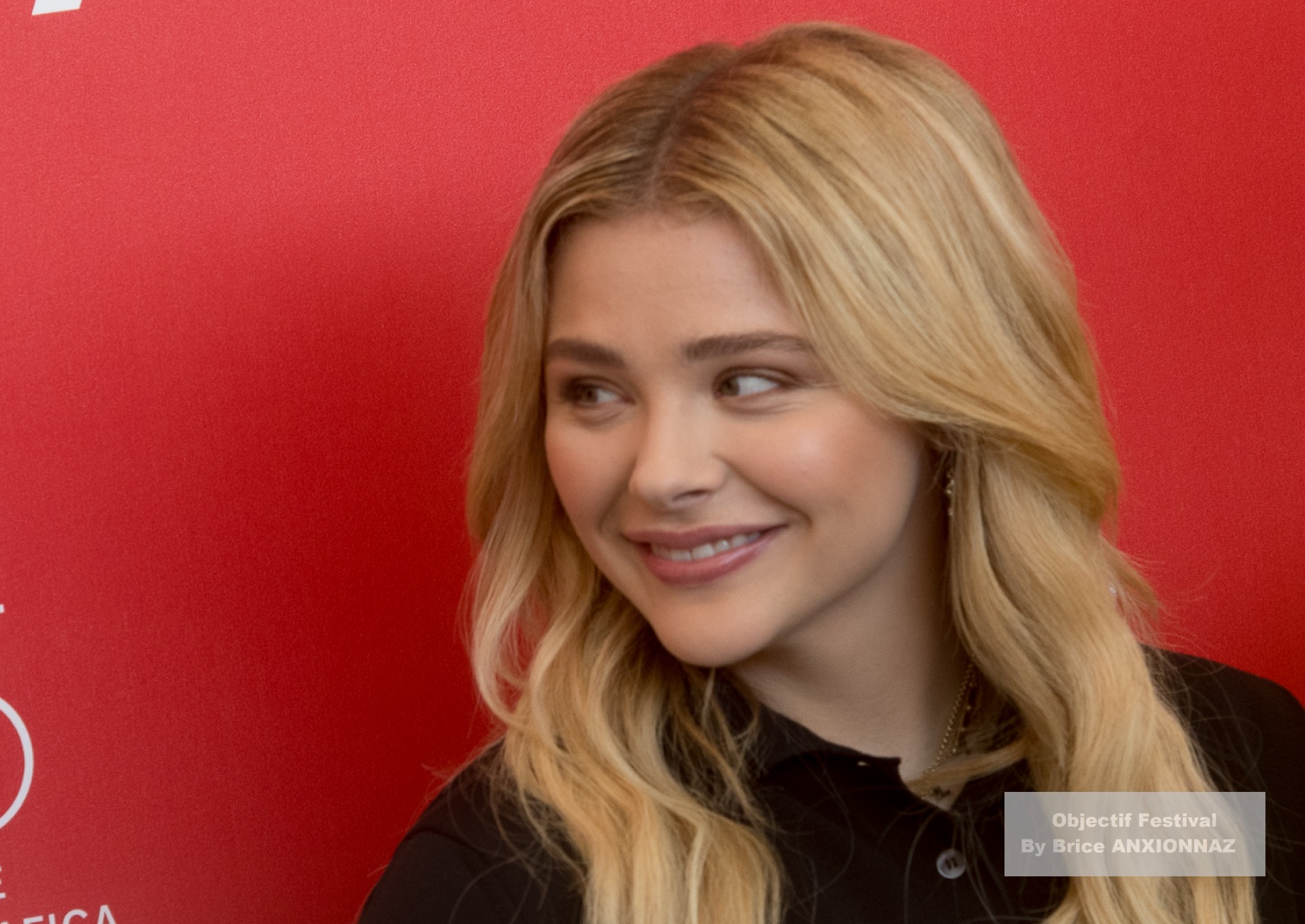 Suspiria — Chloe Grace Moretz — Une personne souriante, vêtue d'un haut noir, se tient devant un fond rouge, créant une ambiance chaleureuse et amicale.