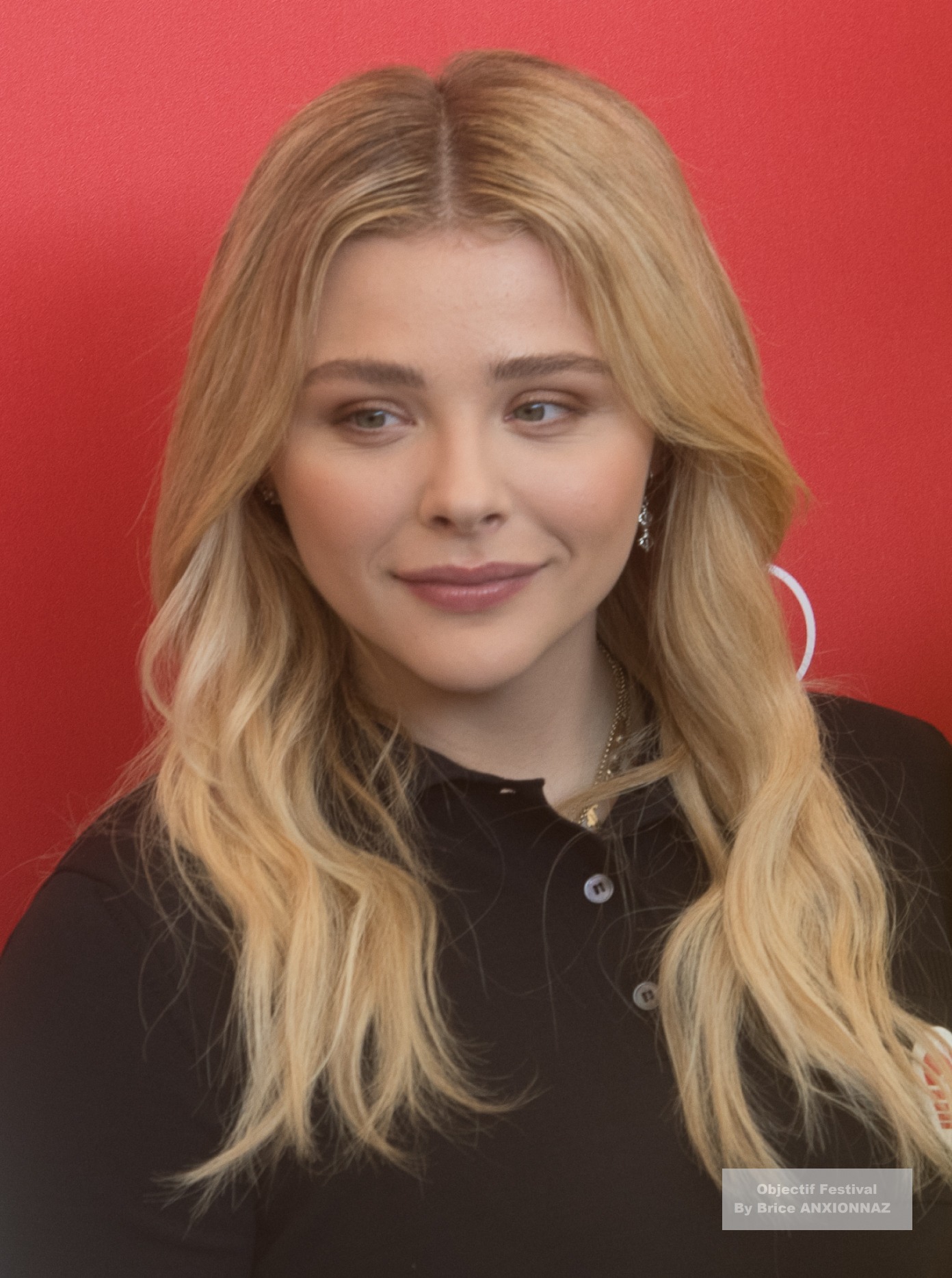 Suspiria — Chloe Grace Moretz — Une personne avec des cheveux blonds et longs porte un haut noir, se tenant devant un fond rouge, affichant une expression sereine.