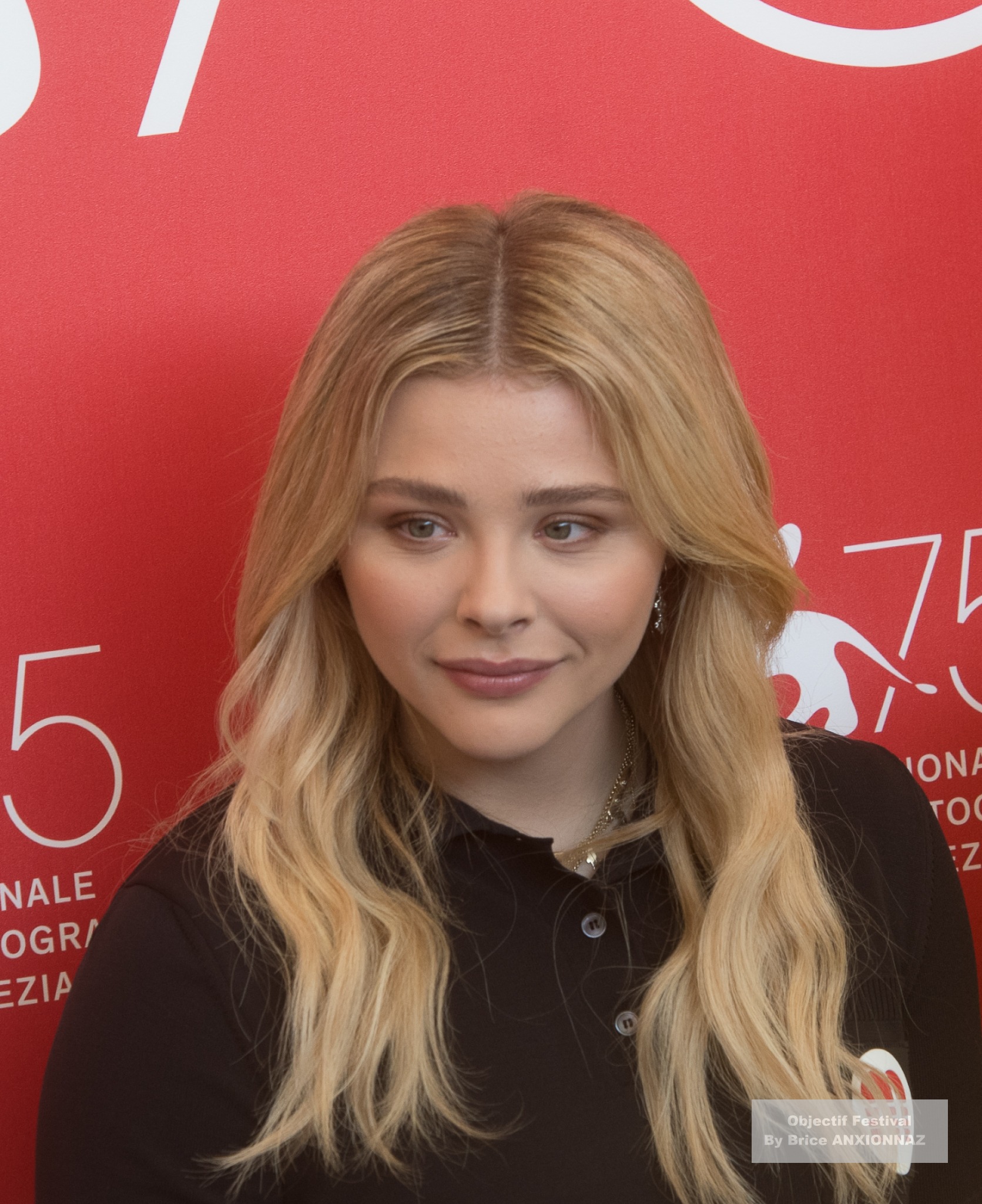 Suspiria — Chloe Grace Moretz — Une personne avec des cheveux longs et blonds porte un polo noir, se tenant devant un fond rouge avec des inscriptions blanches, dégageant une ambiance calme et réfléchie.