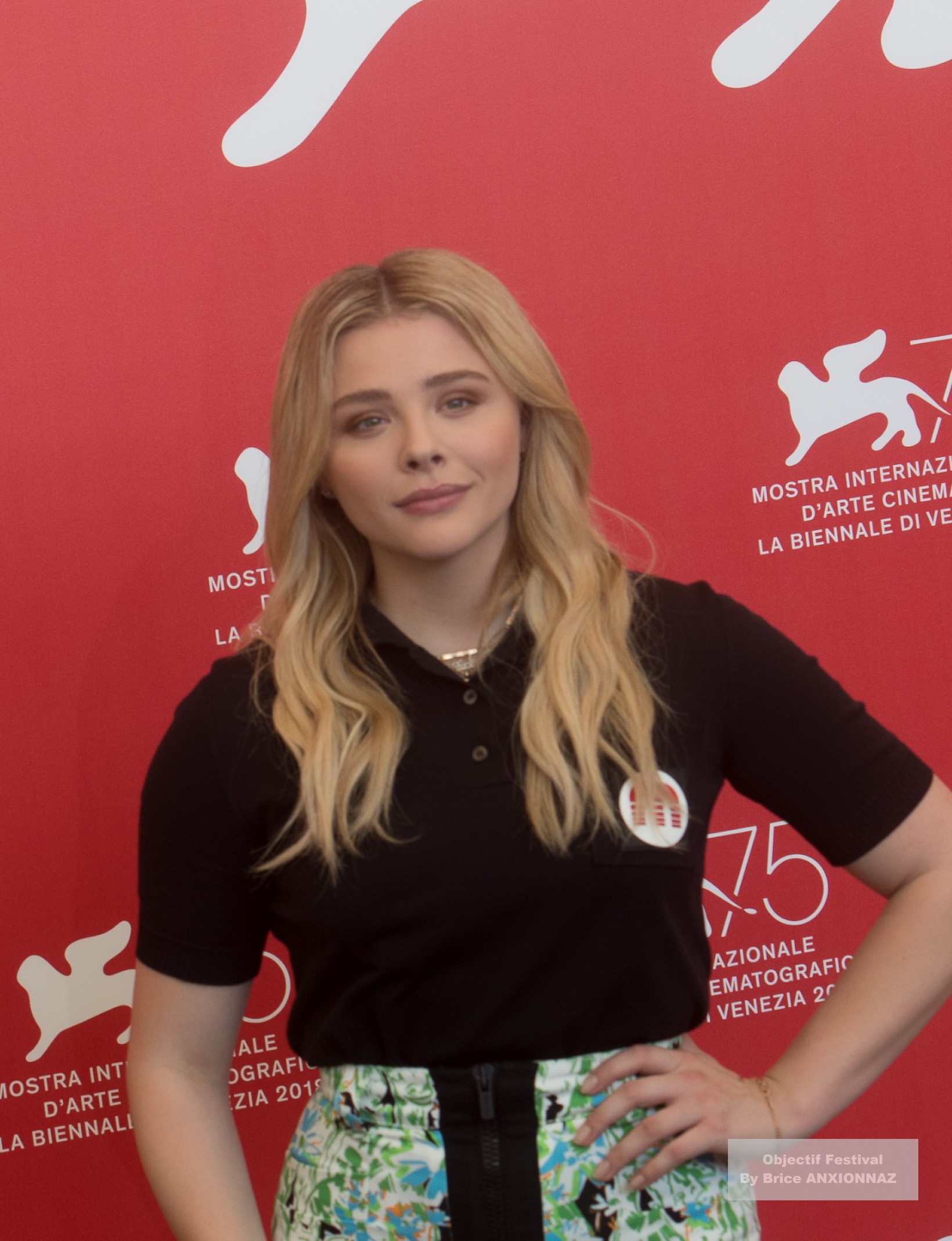 Suspiria — Chloe Grace Moretz — Une personne se tient devant un fond rouge avec des inscriptions blanches, portant un polo noir et une jupe colorée, affichant une expression neutre et posant légèrement de profil.