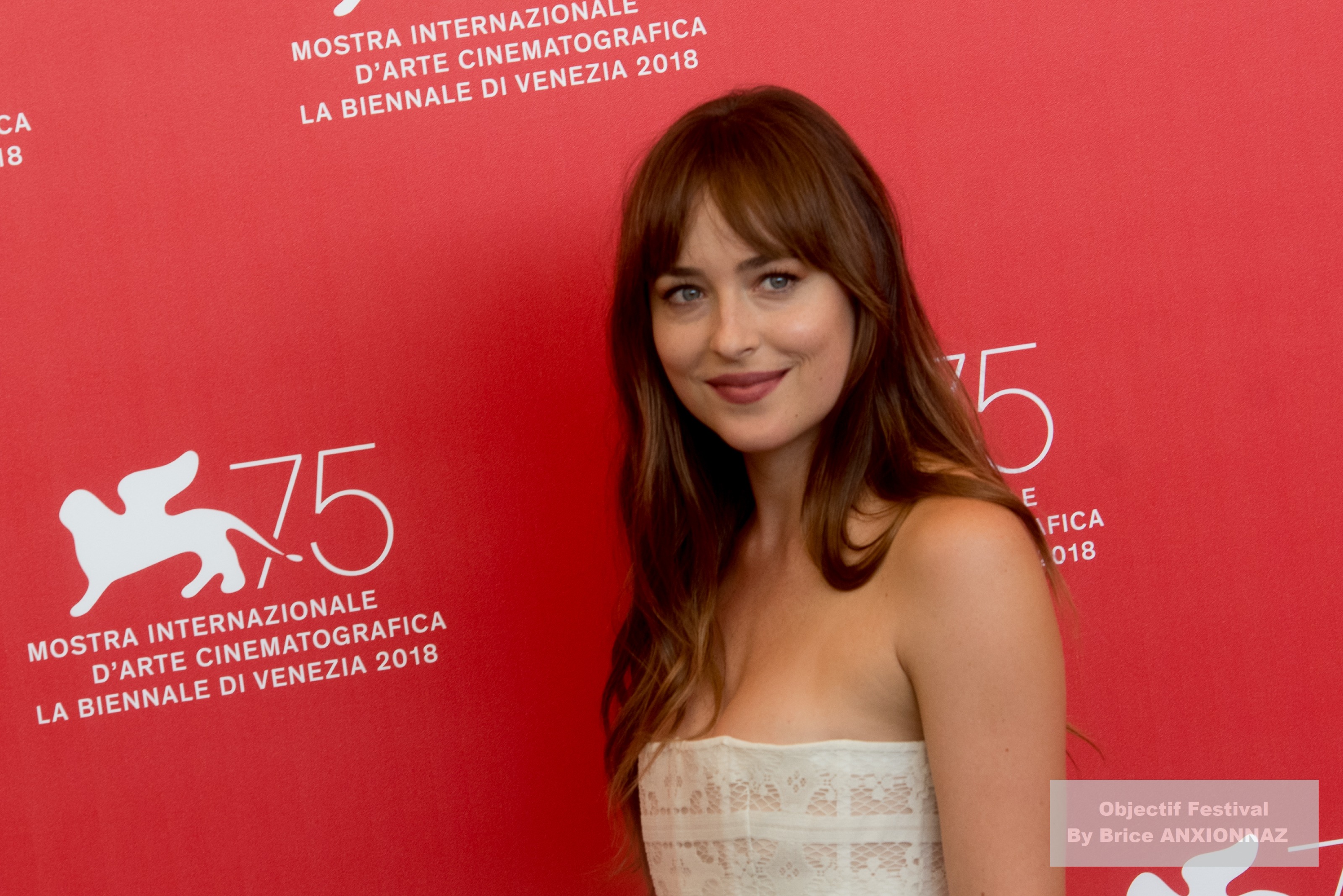 Dakota Johnson — Suspiria — Une personne souriante pose devant un fond rouge avec des logos de festival, portant une robe légère et des cheveux longs, créant une ambiance festive et élégante.