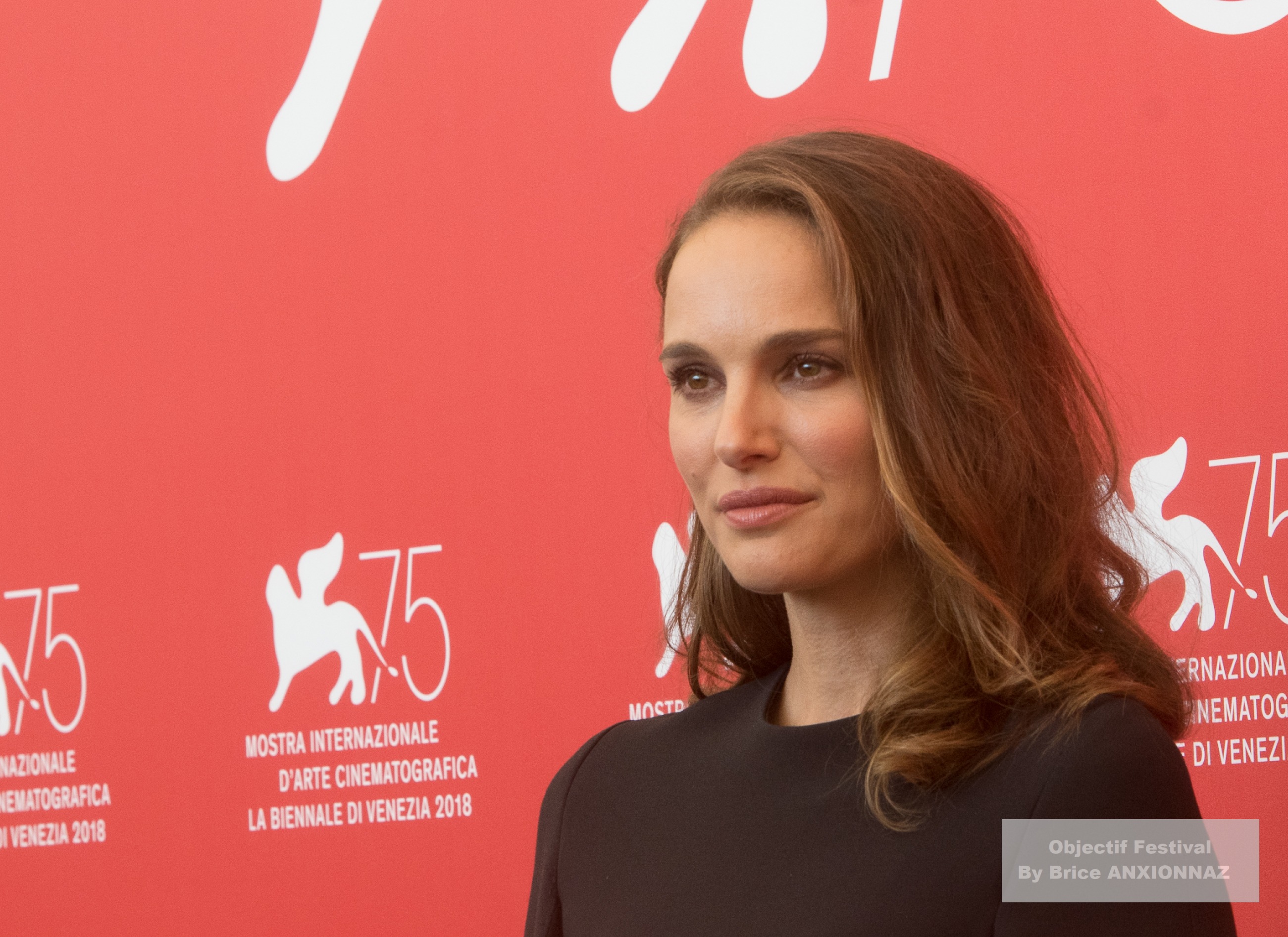 Vox Lux — Natalie Portman — Une personne se tient devant un fond rouge vif avec des inscriptions blanches, portant une tenue sombre et affichant une expression sérieuse, dans une ambiance formelle.