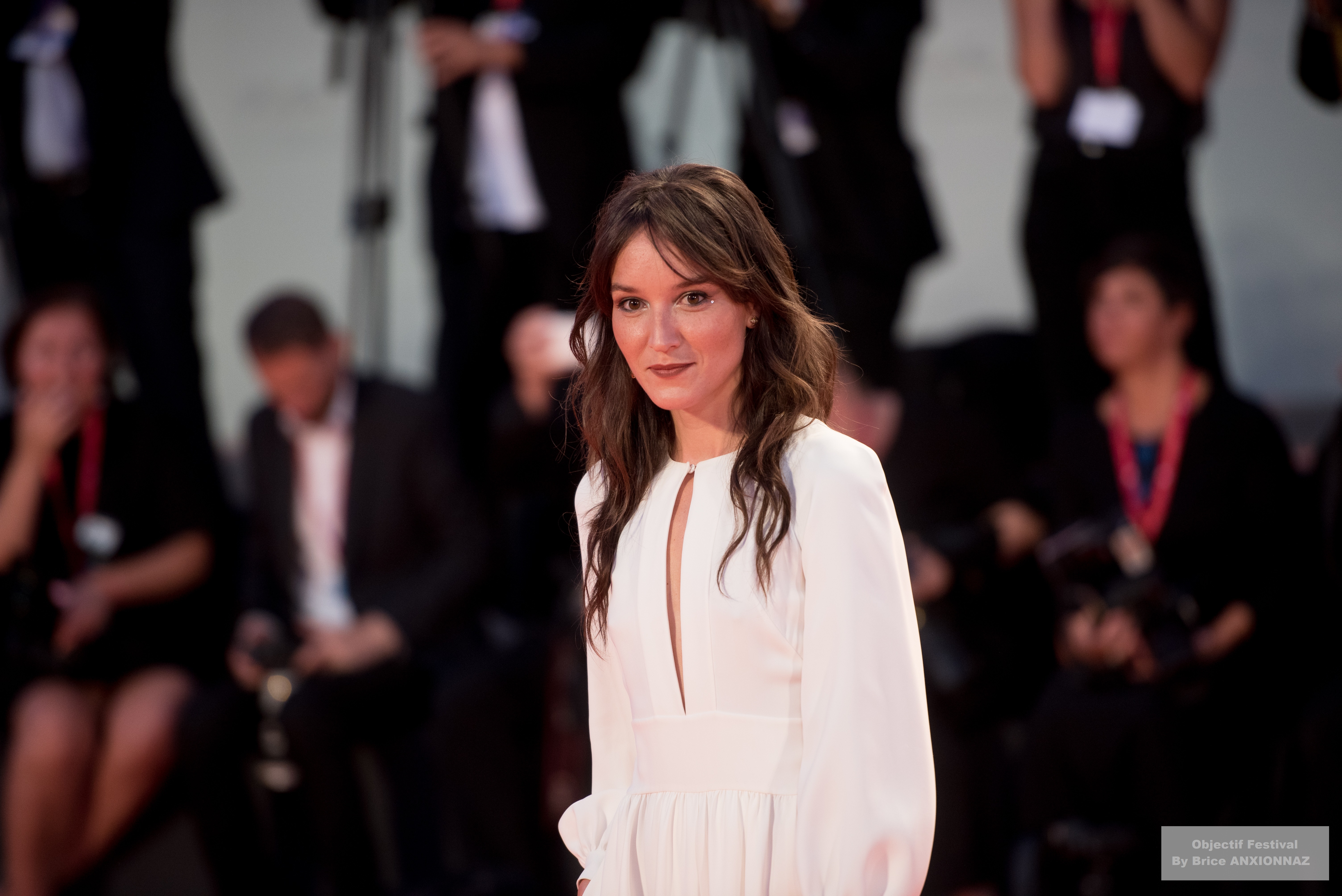 Gloria Mundi — Anais Demoustier — Une personne en robe blanche se tient sur un tapis rouge, entourée de photographes et de spectateurs en arrière-plan, créant une ambiance festive et glamour.