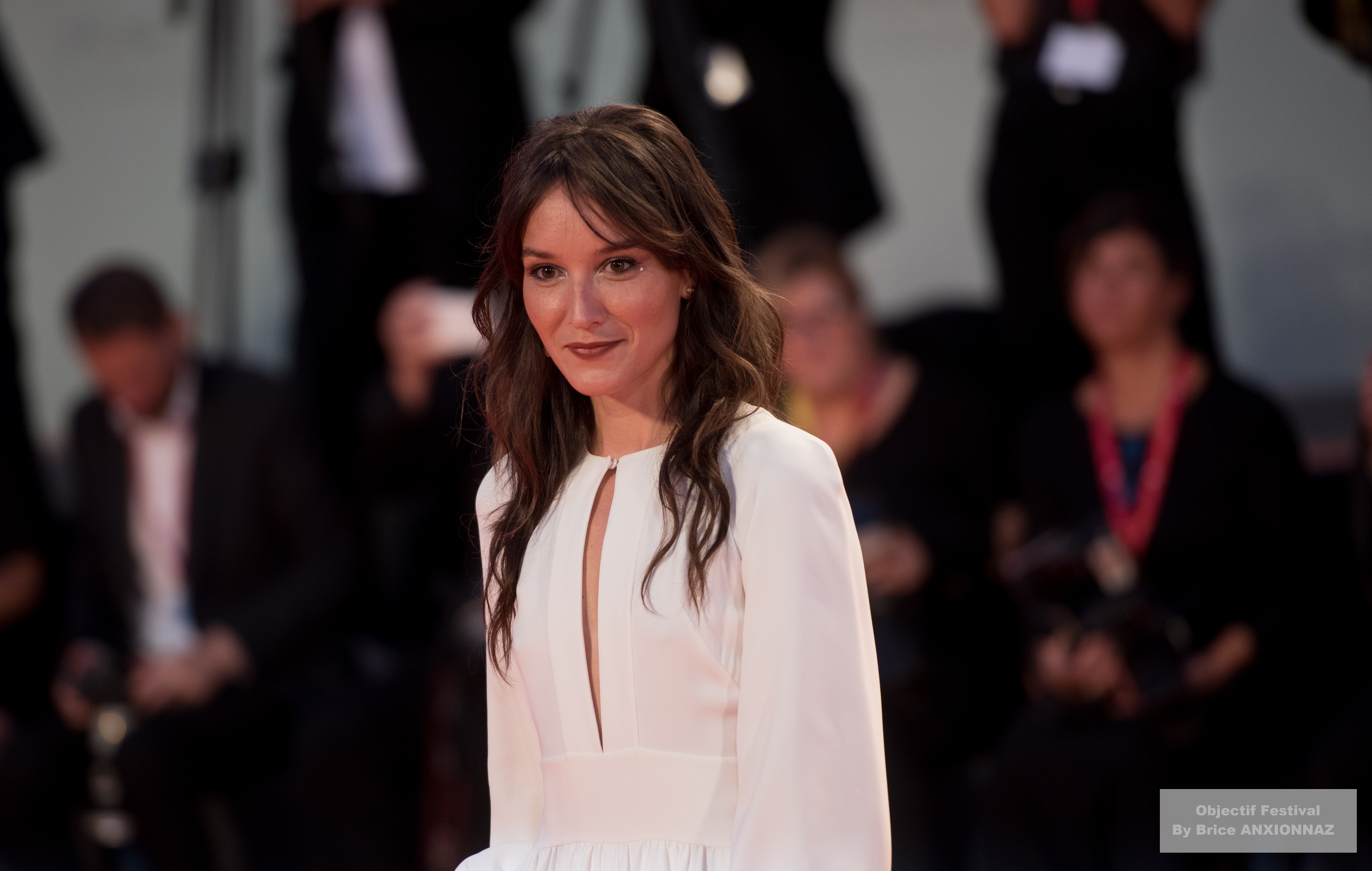 Gloria Mundi — Anais Demoustier — Une personne en robe blanche se tient sur un tapis rouge, souriant, tandis que des photographes et des spectateurs en arrière-plan capturent le moment. L'éclairage est doux et l'ambiance est festive.