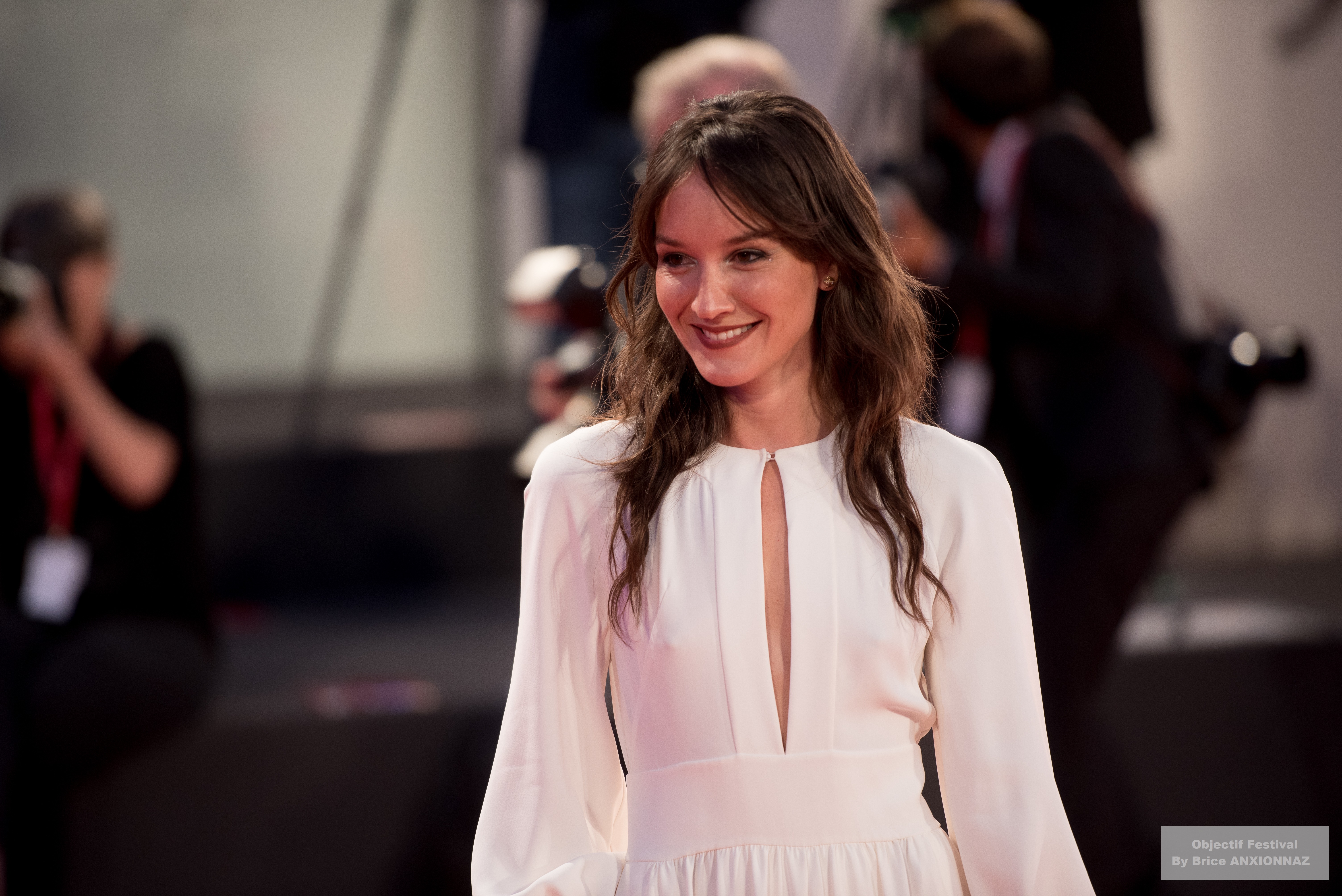 Gloria Mundi — Anais Demoustier — Une personne souriante porte une robe blanche à décolleté, marchant sur un tapis rouge, entourée de photographes en arrière-plan, dans une ambiance festive et lumineuse.
