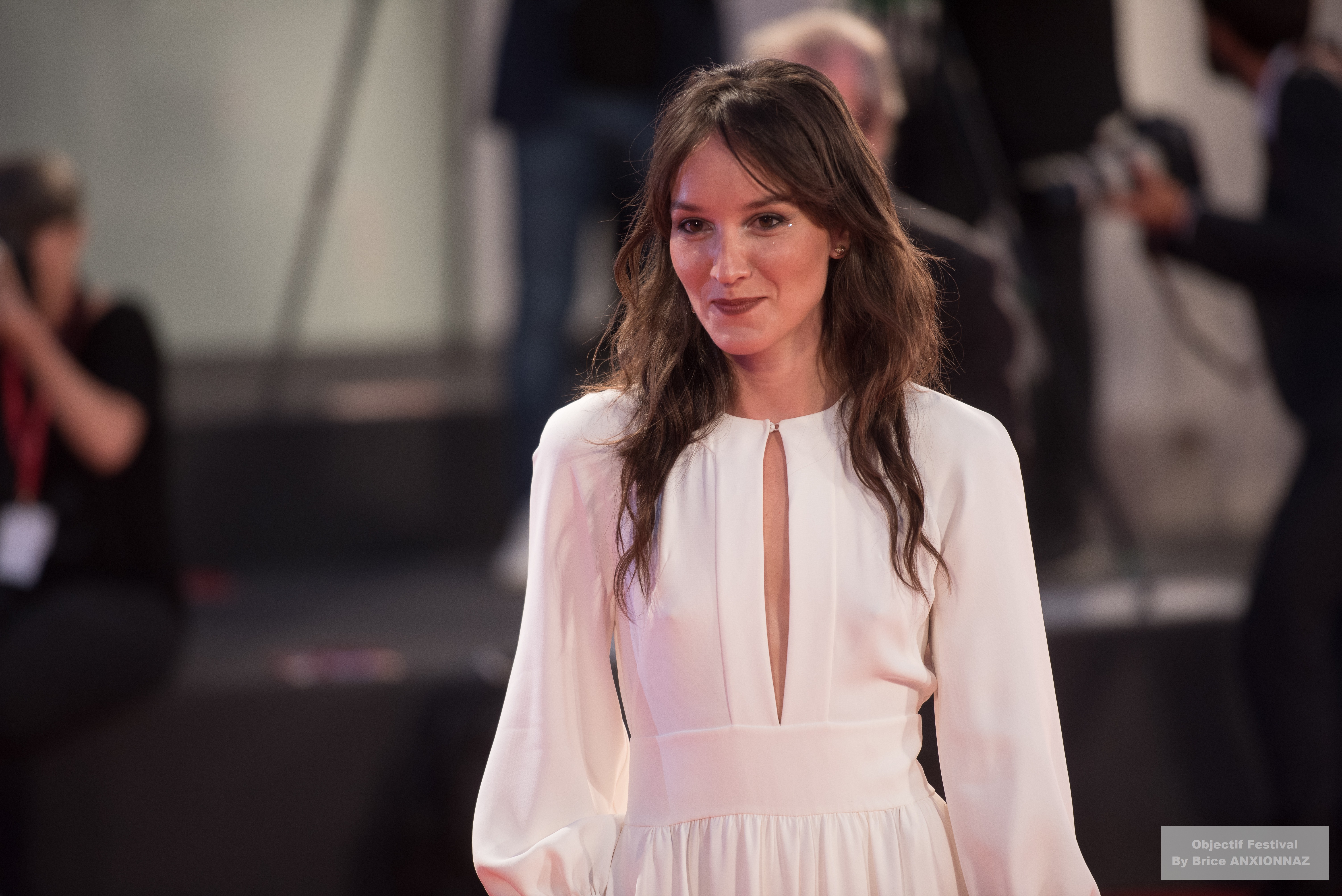 Gloria Mundi — Anais Demoustier — Une personne porte une robe blanche avec un décolleté, souriant sur un tapis rouge, tandis que des photographes capturent l'événement en arrière-plan.