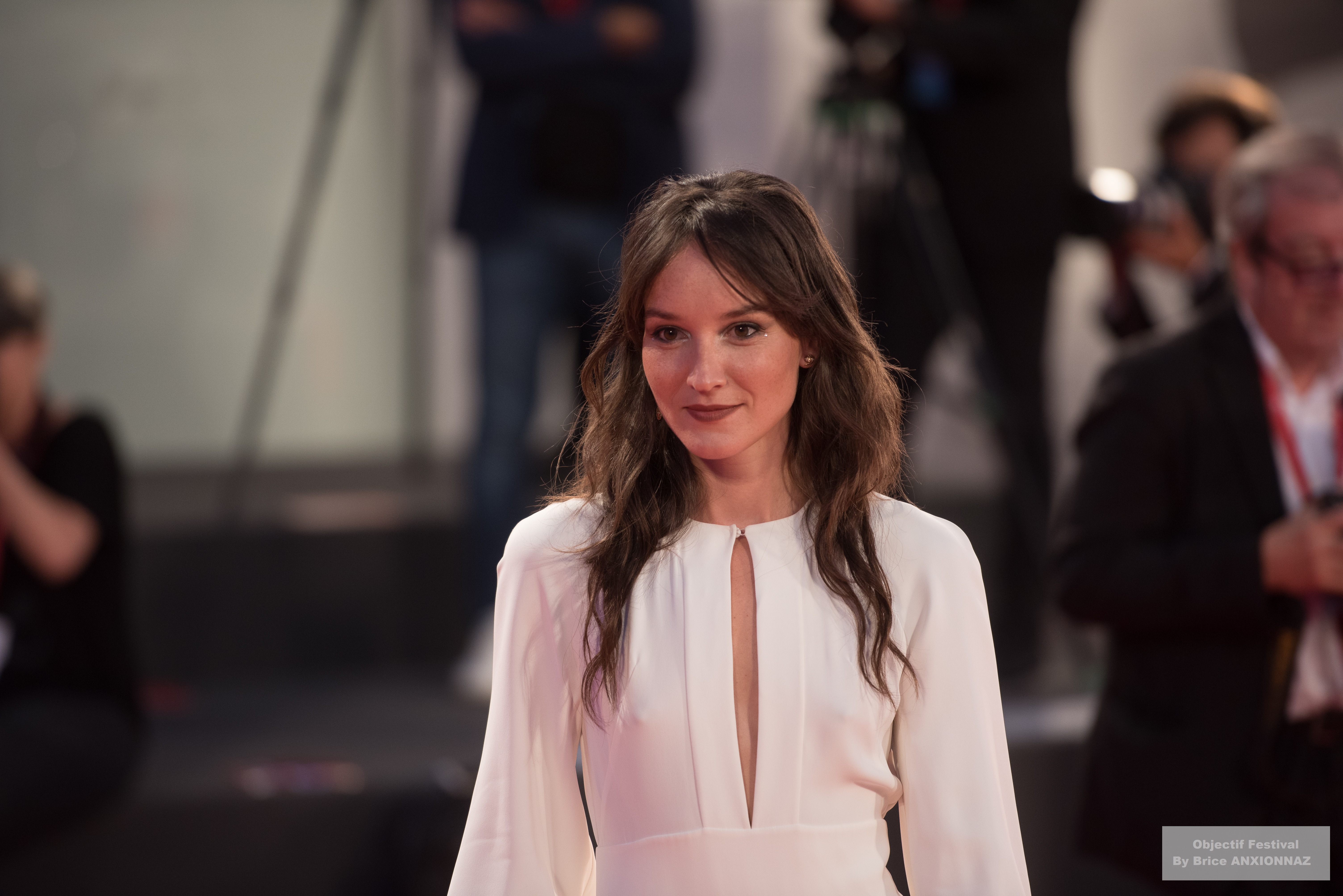 Gloria Mundi — Anais Demoustier — Une personne porte une robe blanche avec une fente, marchant sur un tapis rouge, entourée de photographes et d'une foule floue en arrière-plan, dans une ambiance festive.
