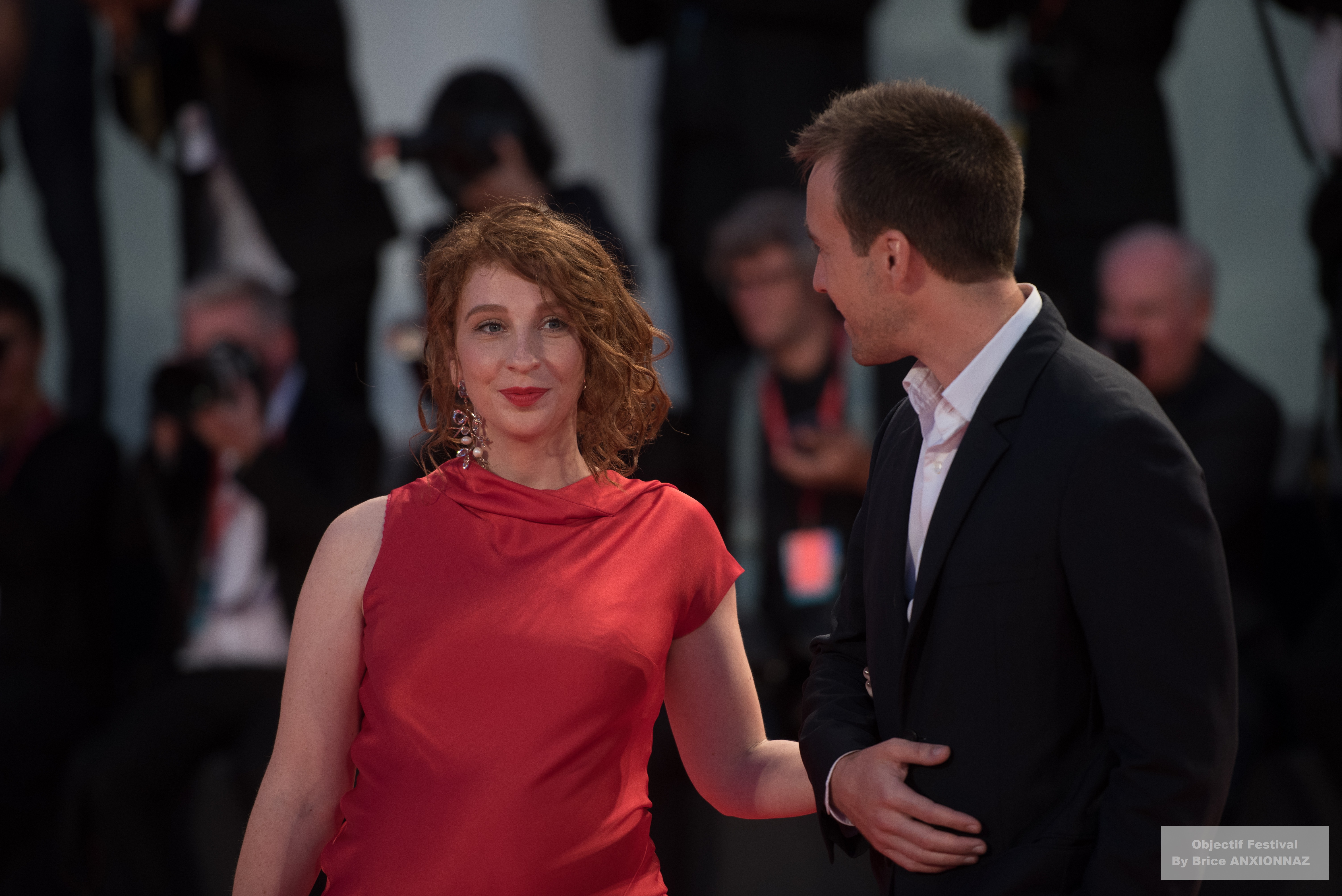Gloria Mundi — Lola Naymark — Une personne en robe rouge sourit tandis qu'une autre, en costume sombre, lui parle, entourés de photographes sur un tapis rouge, créant une ambiance festive et glamour.