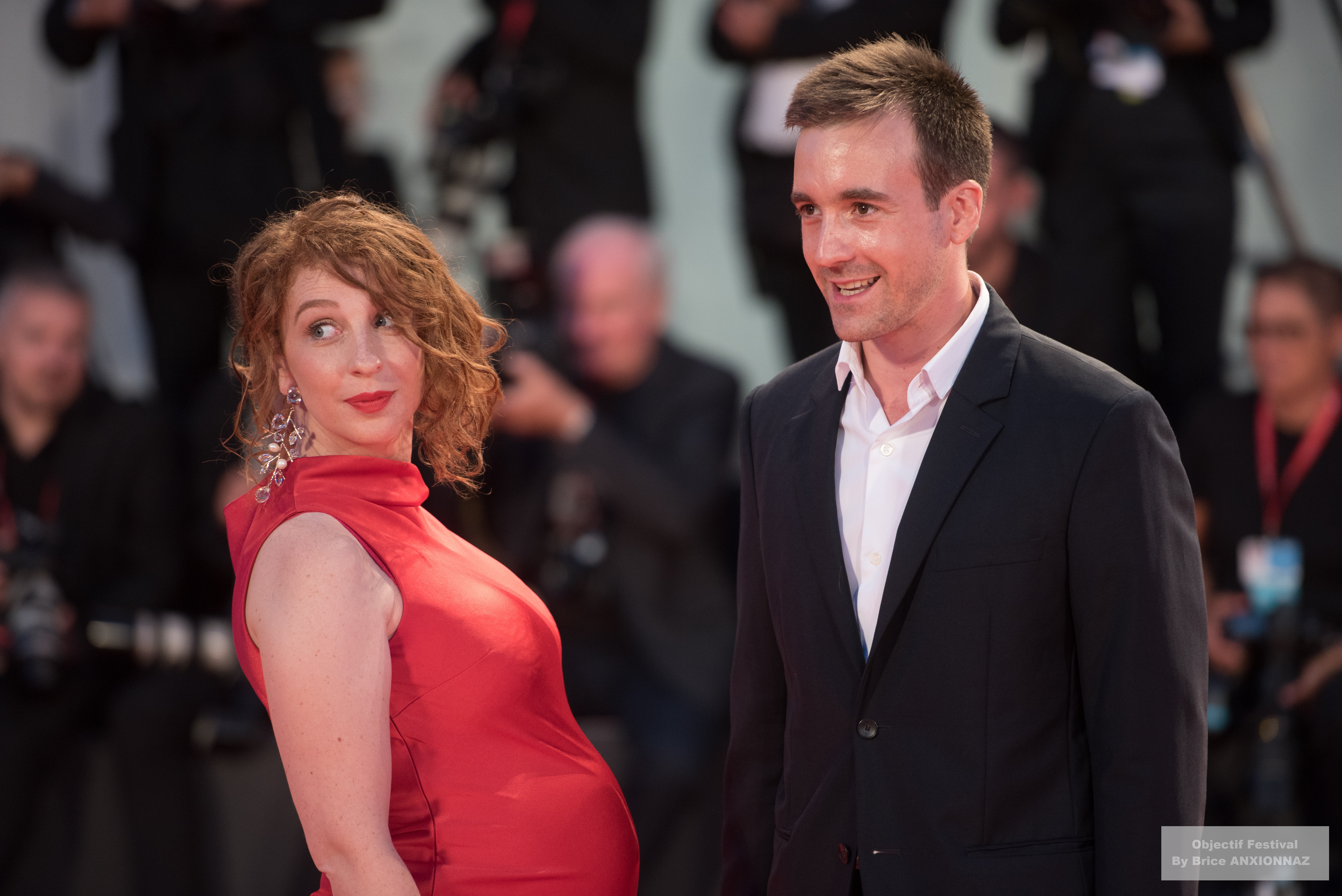 Lola Naymark — Deux personnes posent sur un tapis rouge, l'une en robe rouge et l'autre en costume sombre, avec des photographes en arrière-plan, créant une ambiance festive et glamour. 76Th Mostra Del Cinema, Venise (05/09/2019)