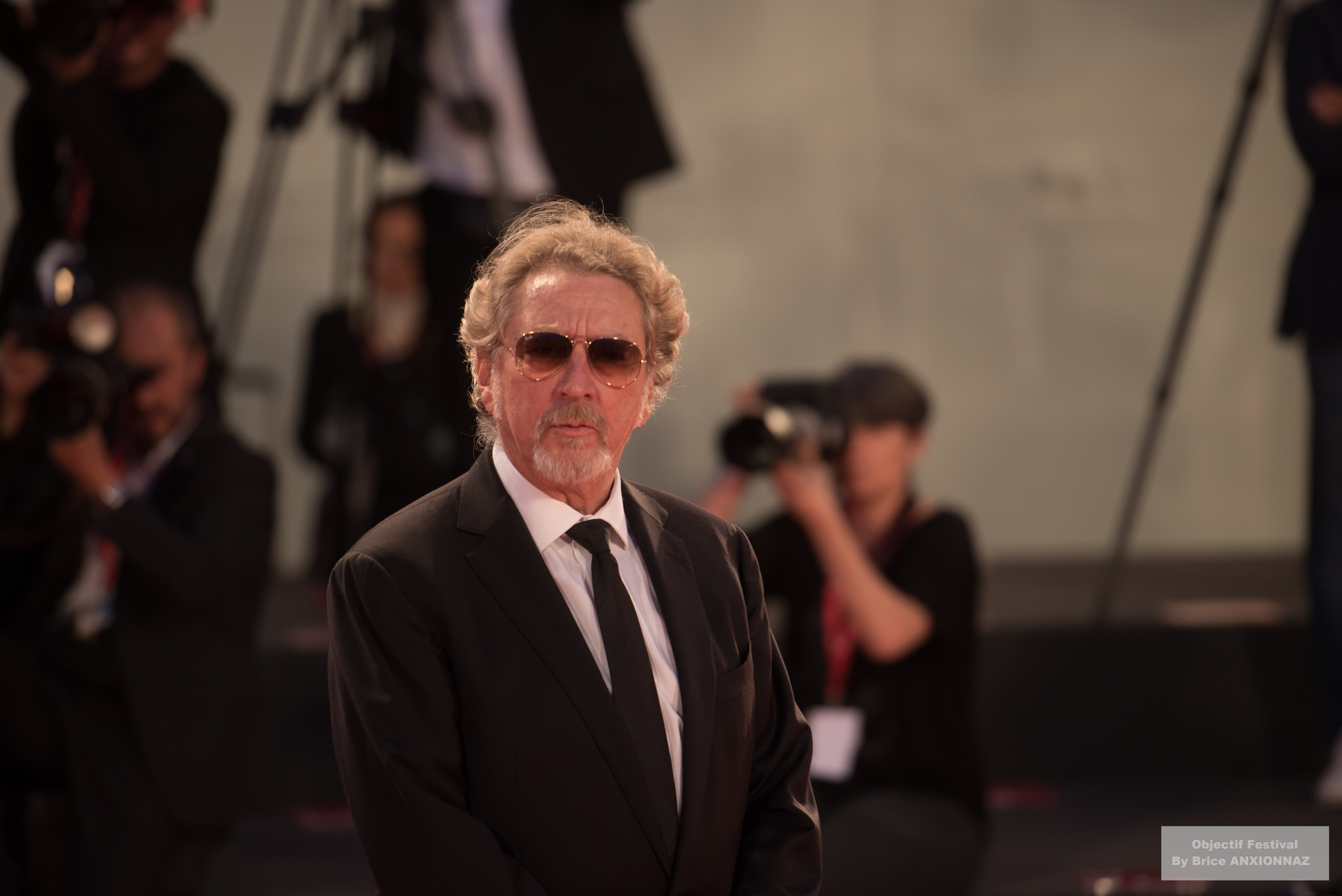 Gloria Mundi — Robert Guediguian — Un homme en costume noir et lunettes de soleil se tient au premier plan, tandis que des photographes en arrière-plan capturent l'événement dans une ambiance lumineuse et festive.