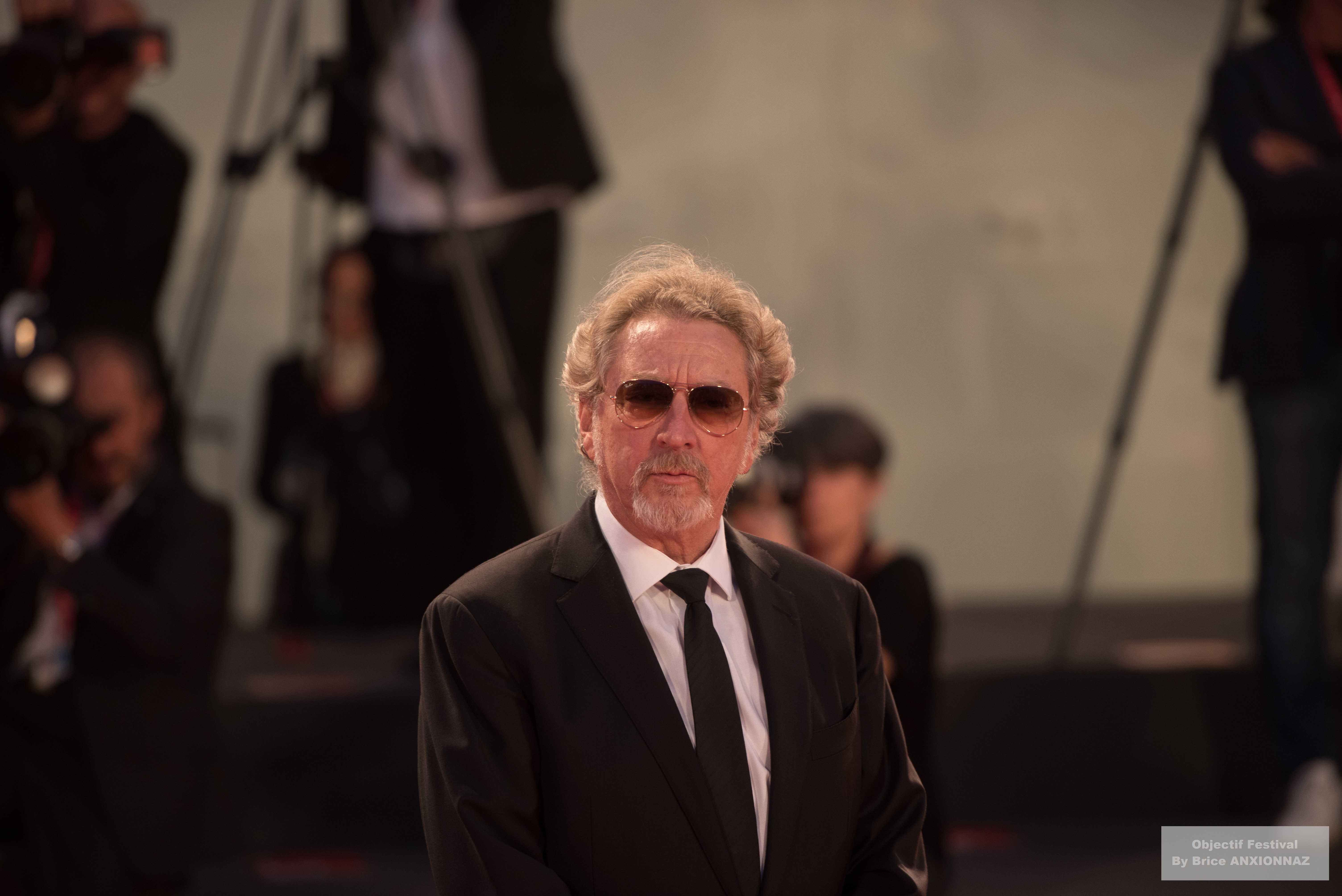 Gloria Mundi — Robert Guediguian — Un homme en costume noir et lunettes de soleil se tient sur un tapis rouge, entouré de photographes, avec une ambiance festive et des lumières vives en arrière-plan.