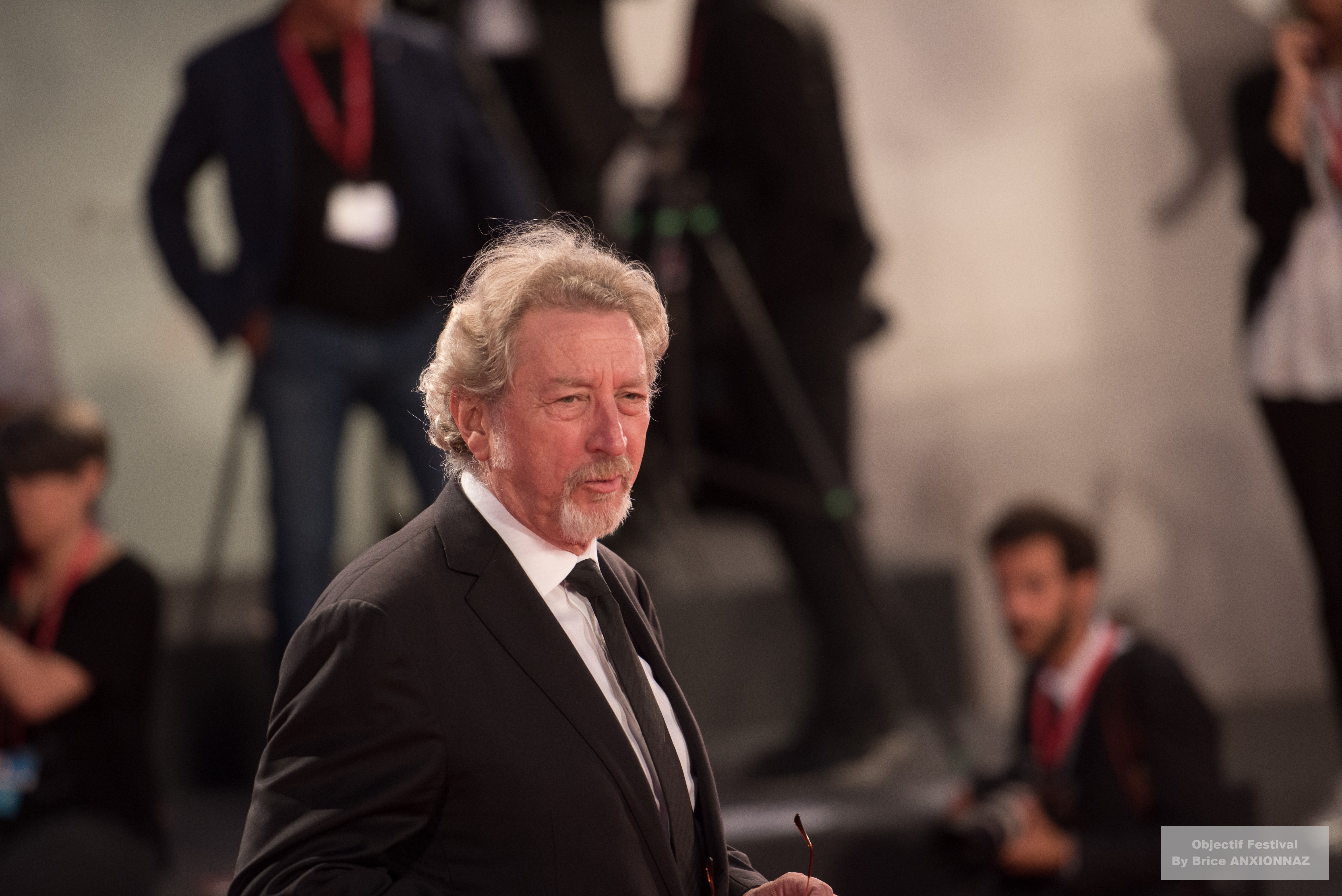 Gloria Mundi — Robert Guediguian — Un homme en costume noir se tient au premier plan, avec des photographes en arrière-plan, dans une ambiance de tapis rouge, sous une lumière douce.